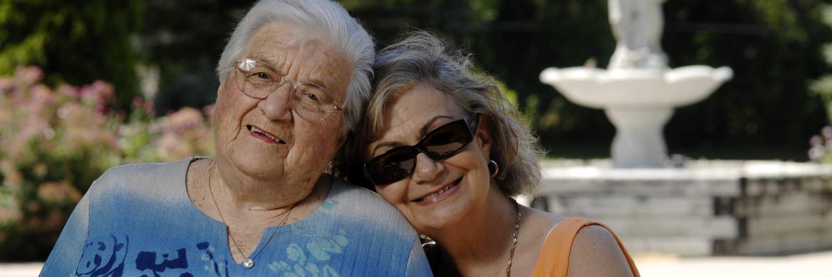 Two older women pose for a photo together. They are sitting outside near a water fountain.