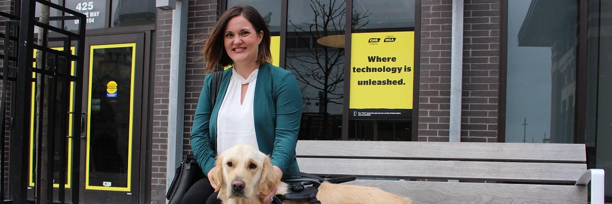 Une femme souriante assise sur un banc avec son chien-guide d'INCA, à l'extérieur d'un édifice d'INCA avec des affiches et des affiches jaunes d'INCA.