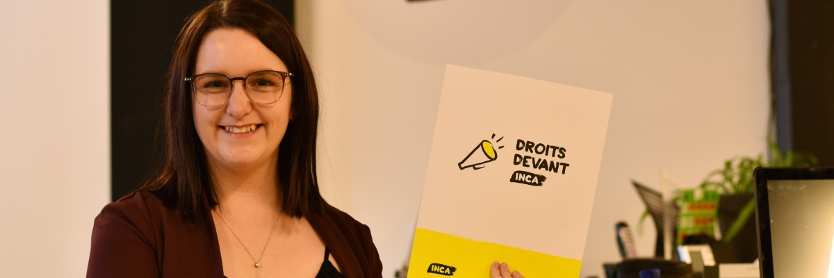 Laurence tient une partie de la trousse, un dossier avec le logo de Droits Devant! d'INCA. En arrière plan sur le mur l'icône d'un porte-voix aux couleurs d'INCA, comme sur le logo.