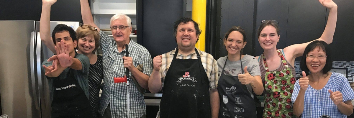 Sept personnes lèvent les bras dans les airs fièrement, derrière un comptoir de cuisine rempli de cupcakes, de glaçage, d'ingrédients et d'ustensiles de cuisine. Un homme tient un canne blanche.