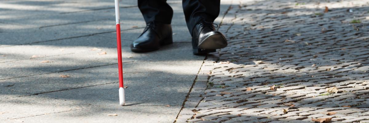 Une canne blanche balaie un trottoir. En arrière-plan, des chaussures d'homme suivent le bout de la canne