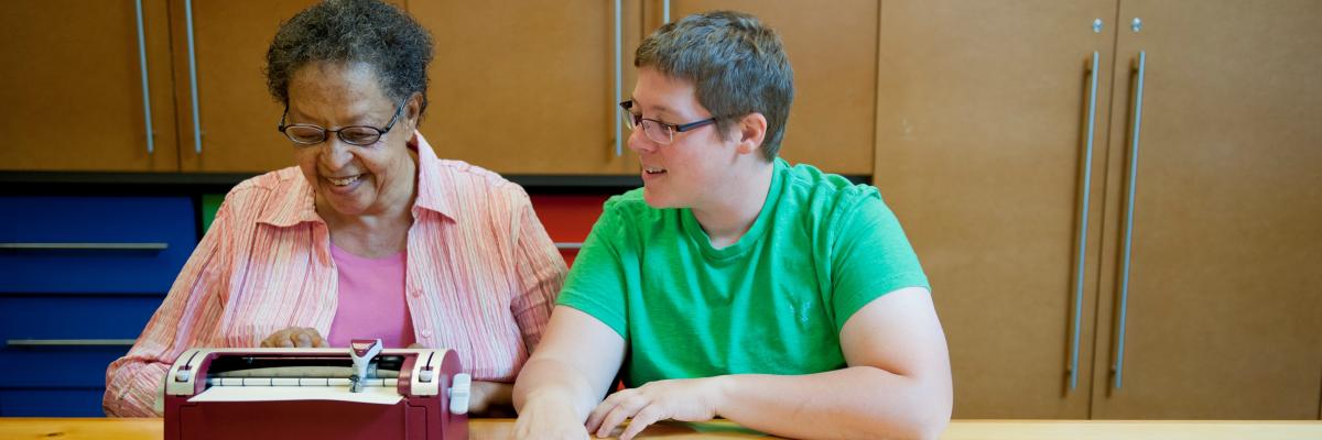 Deux femmes assises à une table sourient pendant que l'une d'elles utilise une machine braille.
