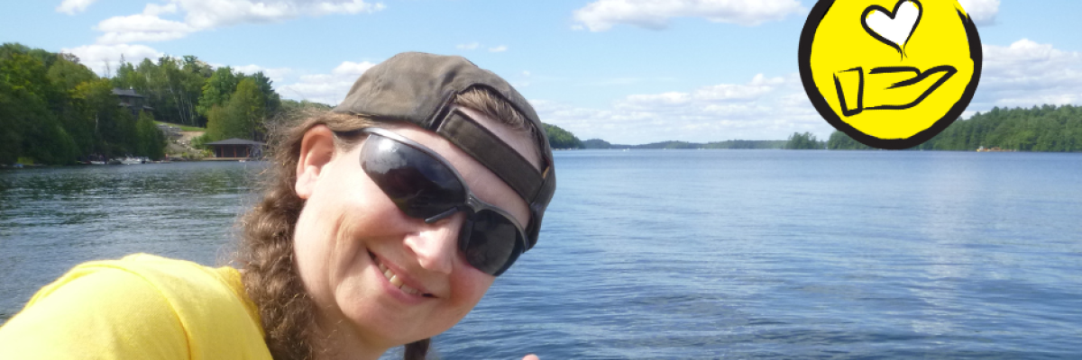 Jessica Bailey pose en face du lac Joe. Elle fait le signe en langue des signes américaine pour "Je t'aime". L'icône d'une main surmontée d'un cœur flottant apparaît dans le coin supérieur droit. 