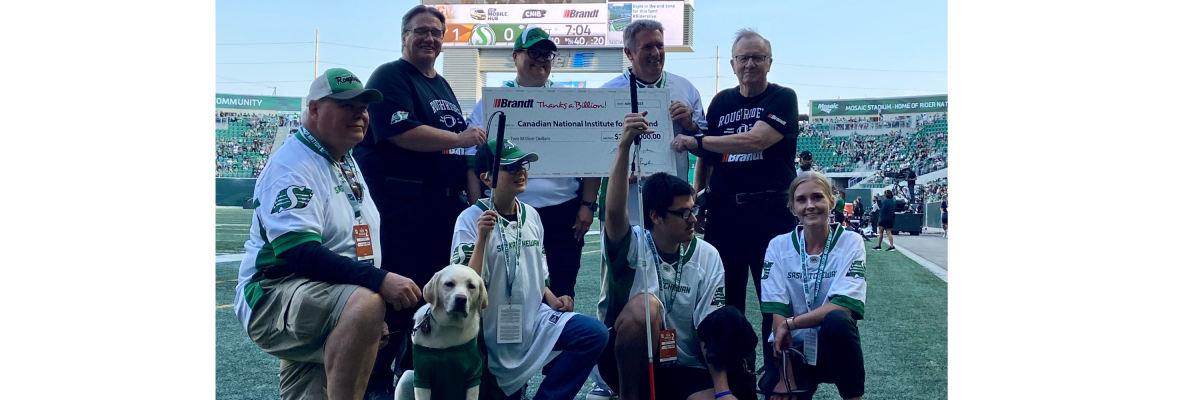 Des représentants d'INCA et du groupe d’entreprises Brandt posent ensemble pour une photo alors qu'ils tiennent le chèque de 2 millions de dollars lors du match des Roughriders de la Saskatchewan. Deux chiens-guides d'INCA portent des chandails verts