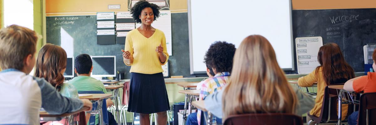 Une jeune femme se tient debout à l'avant d'une salle de classe et s’adresse à un groupe de jeunes élèves. 