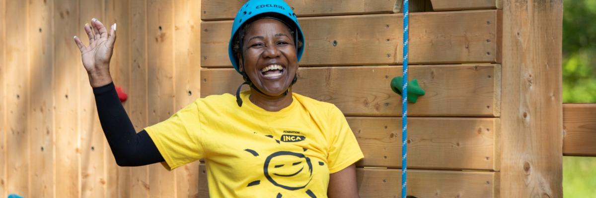 A woman is wearing a yellow CNIB t-shirt and blue helmet at the bottom of the climbing tower. She is smiling with one hand held up.