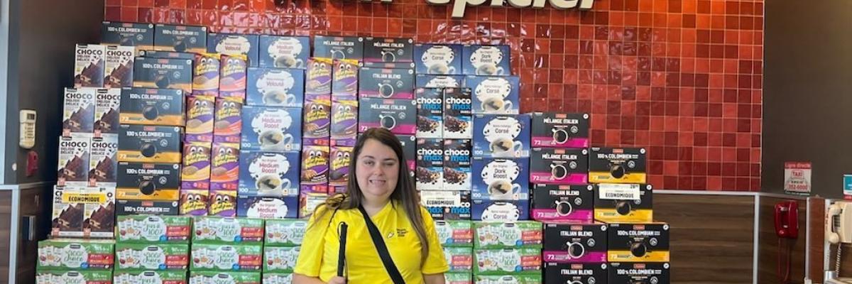 Carolane portant son chandail Quartier Accessible devant l'insigne de l'épicerie Métro avec à la main droite sa canne blanche