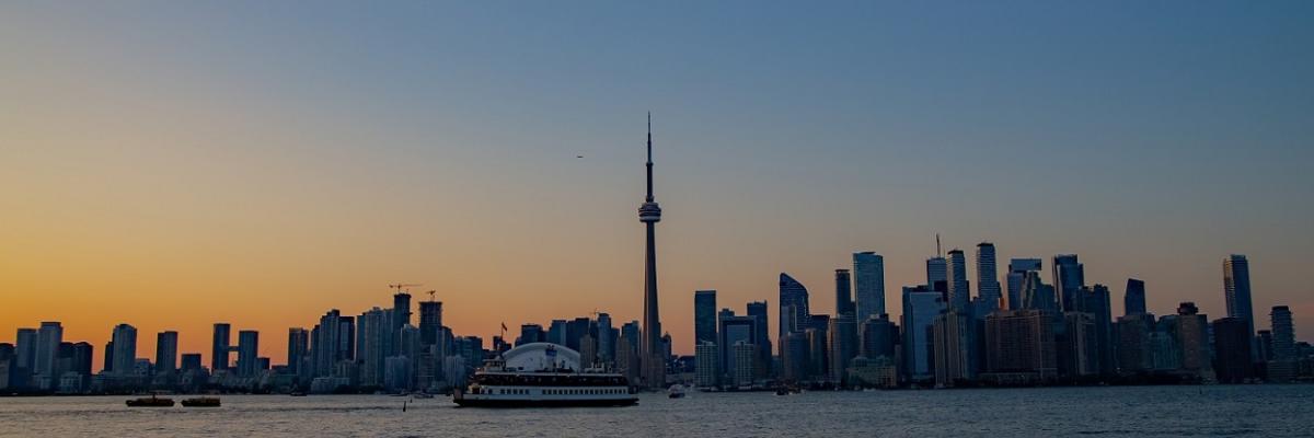 The Toronto skyline at sunset taken from Toronto Island, this is the view many enjoy when on a boat cruise in Toronto harbour. The sky is a darkening rich blue and the sunlight in yellow and gold is framing the buildings and skyline. You can see a ferry crossing. 