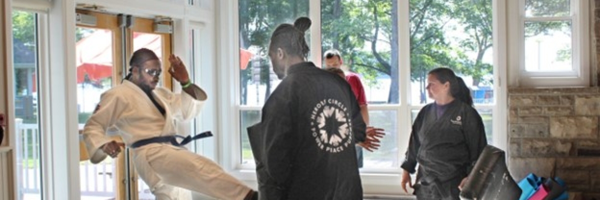 Un jeune homme noir portant un uniforme de karaté blanc et une ceinture noire donne un coup de pied sur un coussin qu'un homme tient devant lui.