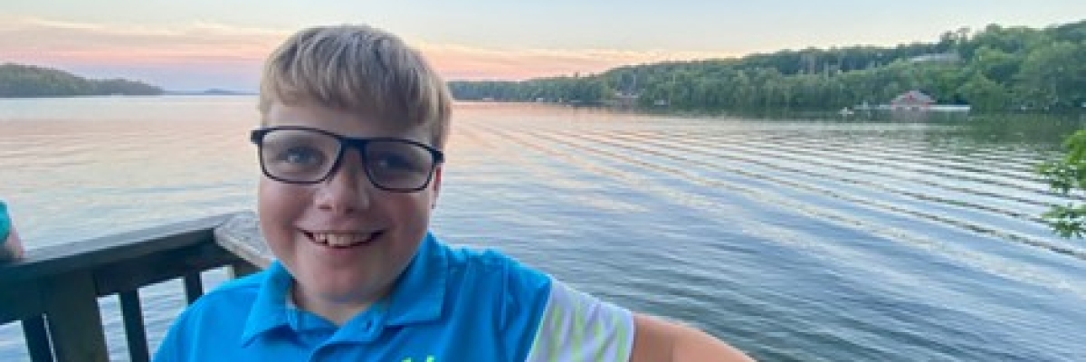 Un jeune garçon portant des lunettes et tenant une canne blanche s'appuie contre une balustrade en bois. Derrière lui, un lac entouré d’arbres à l'horizon.