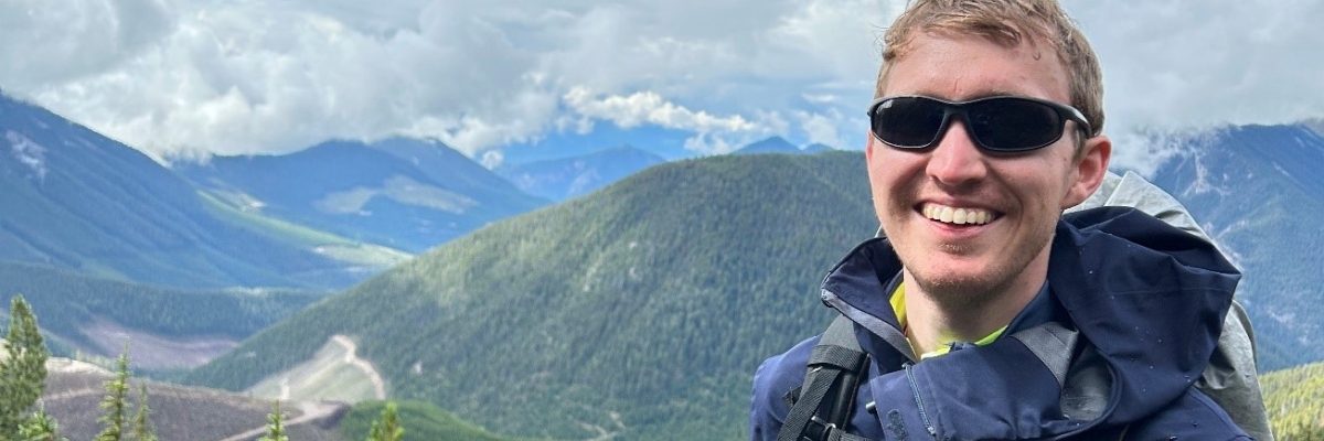 Un jeune homme se tient en plein air, sur une colline et on aperçoit des montagnes en arrière-plan. Il porte des lunettes de soleil noires, une veste bleue, un sac à dos et une canne est attachée à sa ceinture. 