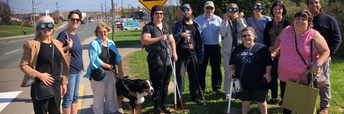 Un grand groupe de personnes souriantes se tient à côté d'un passage pour piétons situé près d'une grande route L'une des personnes a un chien d'aveugle, plusieurs autres portent des lunettes qui simulent différentes expériences de la cécité. 