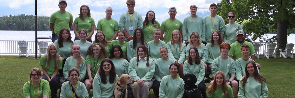 Les membres du personnel d'INCA Lake Joe posent pour une photo de groupe à l'extérieur avec le lac derrière eux. Ils portent tous des t-shirts et des sweat-shirts verts.
