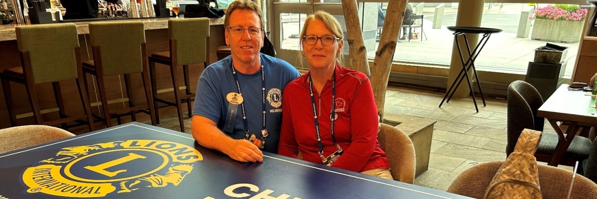 Jerry et Jenn sont assis ensemble derrière une table sur laquelle sont imprimés le logo du Lions International et les mots "Cheers to Kindness".