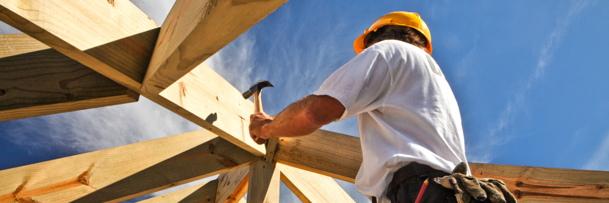 Un ouvrier du bâtiment, coiffé d'un casque de chantier, utilise un marteau pour fixer des poutres en bois, vu d'en bas sur fond de ciel bleu éclatant.