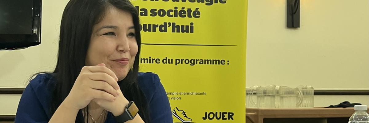 Najla sourit assise, les mains jointe à un table, devant la bannière Changer ce que signifie être aveugle d'INCA.