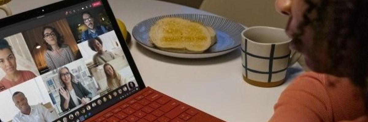 Une femme assise à une table avec un café participe à une rencontre de groupe sur son ordinateur. Les autres participants sont en vidéos.
