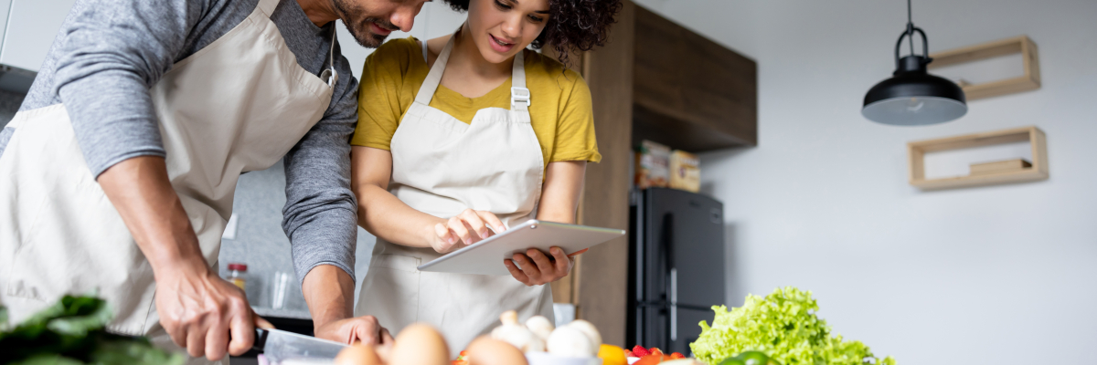 Un jeune homme et une jeune femme portant un tablier, consultent une recette sur une tablette.  Le jeune homme coupe une partie des ingrédients qui sont devant eux.