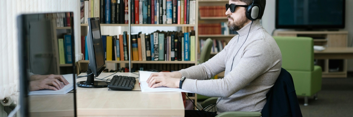 Un homme aveugle avec des lunettes de soleil et un casque est en train de travailler sur son ordinateur.