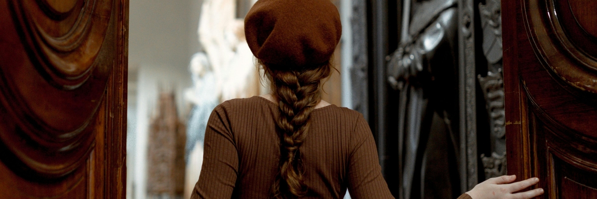 Une femme coiffée d’un béret brun entre dans une salle de musée ornée de sculptures en poussant une grande porte en bois.