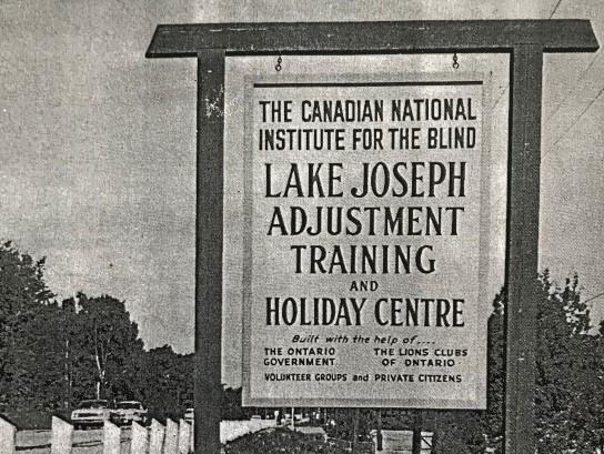 Une photo en noir et blanc du panneau de l’entrée du Adjustment Training and Holiday Centre de l'Institut national canadien pour les aveugles au camp Lake Joseph en 1965.