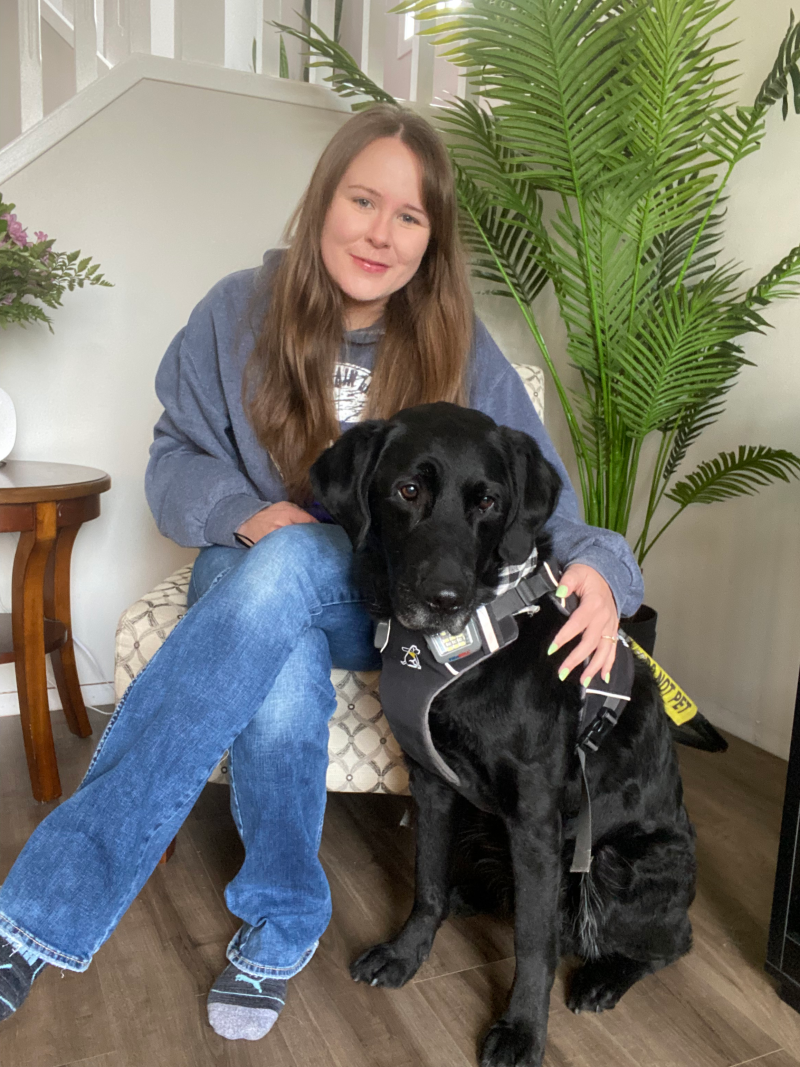 Taylor, une jeune femme aux longs cheveux bruns, est assise sur une chaise, le bras autour de son chien-guide Wallace, un labrador noir portant un harnais d'INCA, qui est assis à ses côtés.