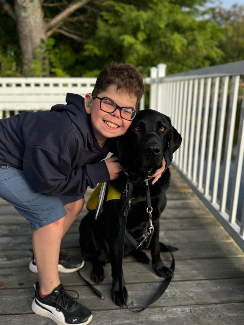 Kaiden, un jeune garçon, est accroupi et enlace de ses bras un labrador noir, Flinn.
