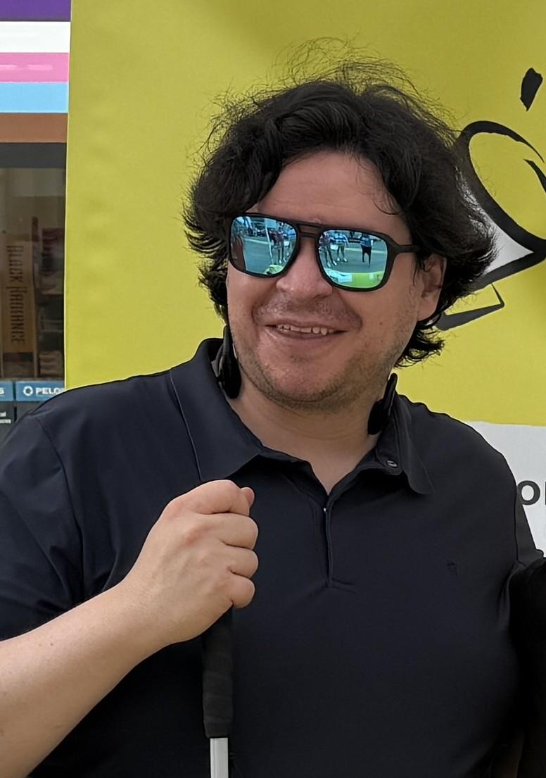 Andres porte des lunettes soleil et tient sa canne. Il est à l'extérieur et sourit.