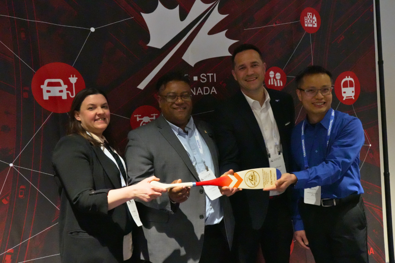 Quatre membres de l'équipe du projet pilote « Zones de construction de trottoirs accessibles aux personnes aveugles et à basse vision » posent ensemble lors d'un événement, souriants et tenant leur prix, une pagaie en bois sur laquelle sont gravées les informations relatives à cette distinction. Mary Ann, à l'extrême gauche, tient une extrémité de la pagaie tandis que les autres membres de l'équipe se rassemblent autour d'elle.