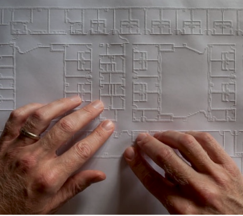 Deux mains parcourent un grand plan architectural tactile. Des lignes en relief délimitent les pièces, les corridors, les murs et d’autres éléments de la disposition du bâtiment.