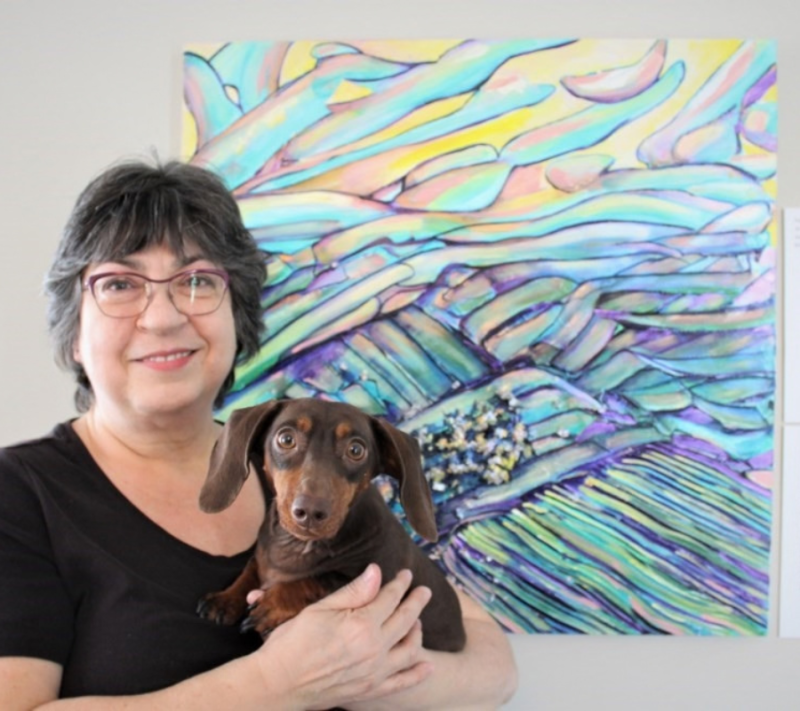 Robyn Rennie pose pour une photo devant son œuvre d’art colorée, à côté d’un code QR imprimé numériquement. Elle tient dans ses bras un petit chien de couleur brun chocolat.
