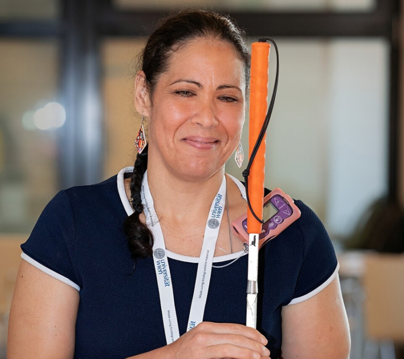 Dr. Wanda Díaz-Merced smiles while holding an orange-handled white cane. She wears a Universidad de Valencia lanyard.