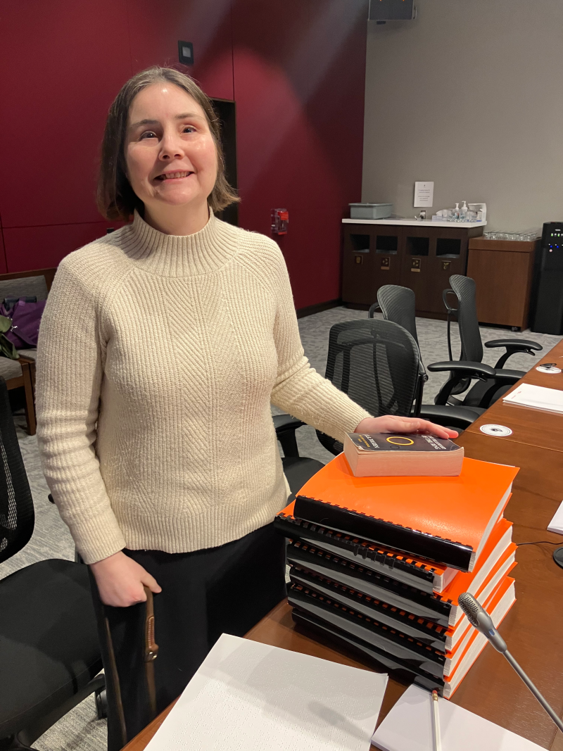 Jen Goulden sourit, debout devant un bureau dans une salle de réunion du Comité sénatorial. Sur le bureau, elle tient son exemplaire en braille du roman « La Communauté de l’Anneau », composé de six volumes épais et lourds. Au sommet de la pile se trouve une version de poche standard du livre, minuscule en comparaison. Ces accessoires ont été utilisés pour illustrer les coûts élevés liés à l’expédition de documents en braille.
