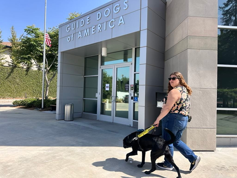 Chelsea Masson se promène avec son chien-guide noir, Rango, devant le bâtiment de Guide Dogs of America par une journée ensoleillée. Chelsea porte un haut sans manches à fleurs, un jean bleu et des lunettes de soleil, tout en tenant le harnais de Rango alors qu’ils passent devant l’entrée.
