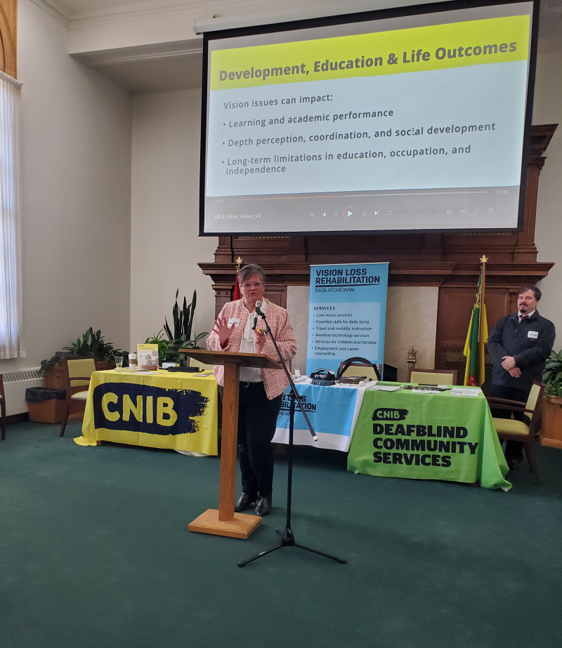 Christall Beaudry, responsable de l'inclusion à INCA, se tient devant un podium et un micro pour prononcer son discours. Derrière elle, on aperçoit des tables arborant les logos d'INCA, du SCS et du VLR, ainsi qu'un diaporama projeté sur grand écran.