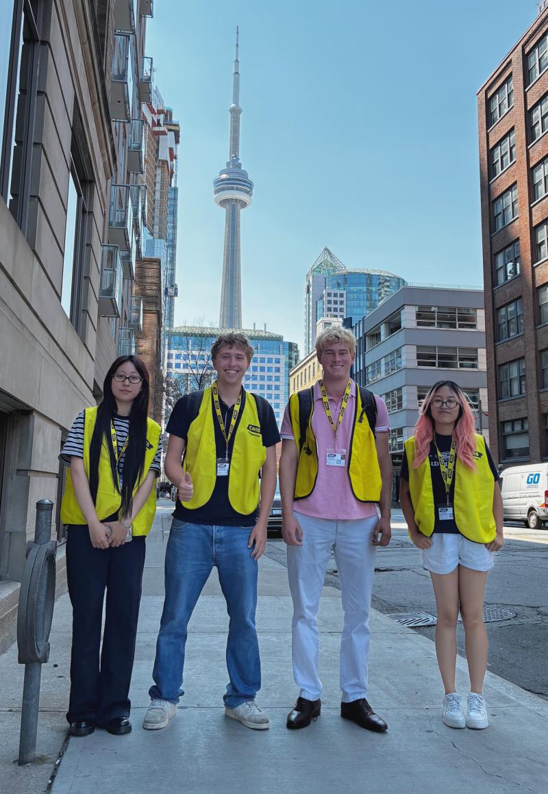 Quatre collecteurs de fonds, vêtus de gilets jaunes d'INCA et portant des cordons d'identification, se tiennent sur un trottoir, la tour CN en arrière-plan. 