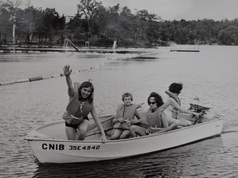 Une photo en noir et blanc des années 1960 de quatre enfants au teint clair portant des gilets de sauvetage et assis dans un bateau sur un lac. Le nom « CNIB » est peint sur le flanc du bateau.