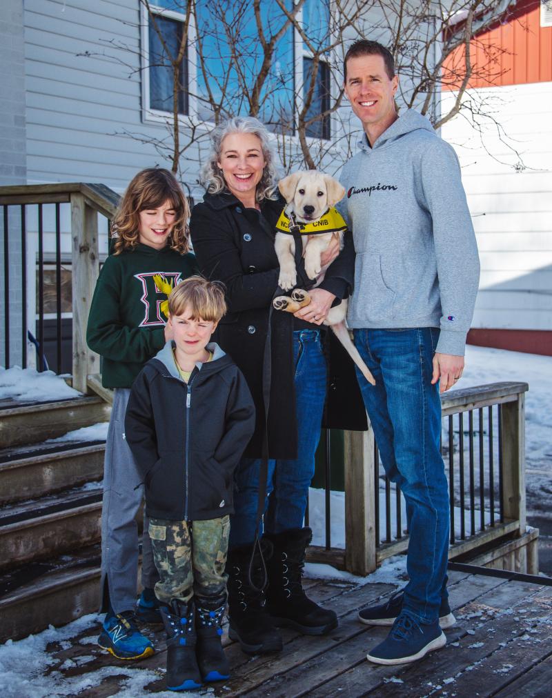 Une famille composée d'une mère, d'un père et de deux jeunes enfants pose devant le porche de leur maison. La mère, qui se trouve au centre, tient dans ses bras un chiot golden retriever. Le chiot porte un gilet jaune du CNIB pour chiens-guides. Il y a de la neige au sol.