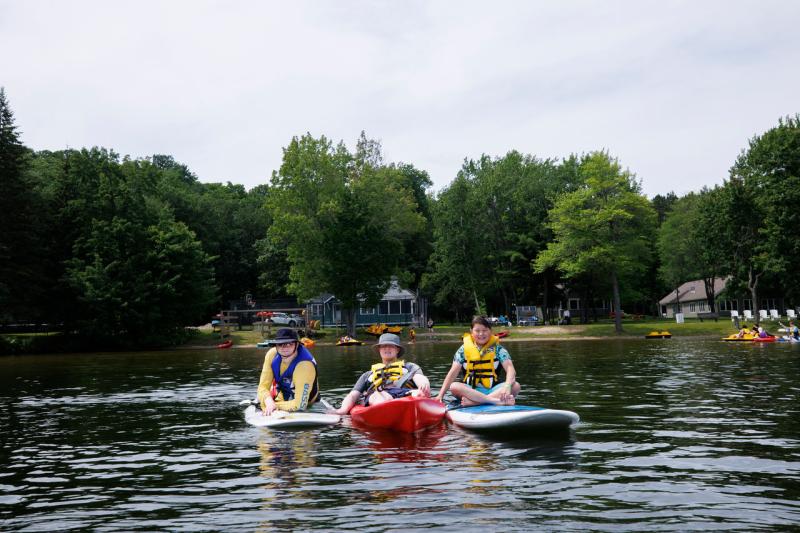 Trois jeunes au teint clair sont sur le lac. Deux d'entre eux sont assis sur des planches à pagaie et le troisième est assis dans un kayak.