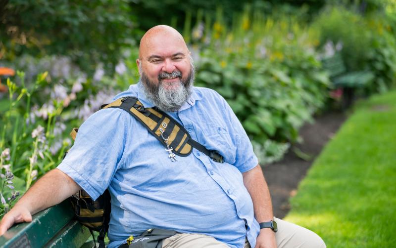 Jason sits on a park bench in front of a small garden, smiling. He is wearing a light blue collared t-shirt, beige pants, and a burnt orange bag