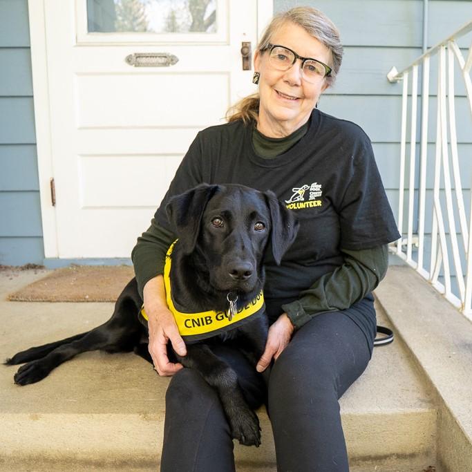Une femme vêtue d'un t-shirt du programme des chiens-guides bénévoles d'INCA est assise sous le porche d'une maison, un chien-guide d'INCA posé sur ses jambes. Le chien est un labrador noir qui porte un gilet jaune de l'INCA.