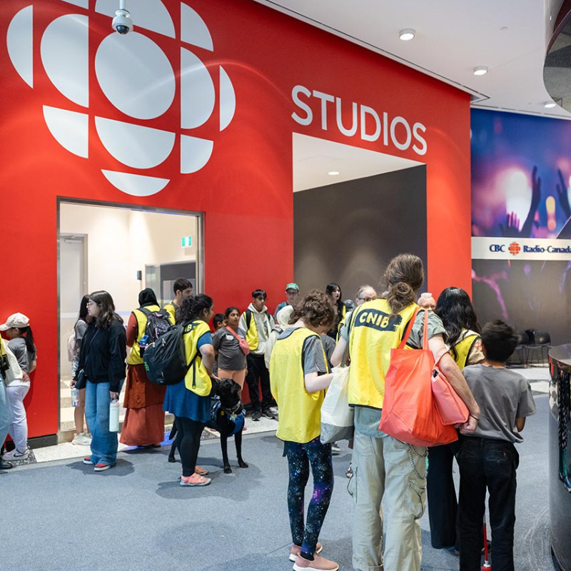 At the Canadian Broadcasting Centre, a large group of young campers and CNIB volunteers gather in an office studio area. Painted on the wall behind them is the CBC logo and the text, “studios.”  