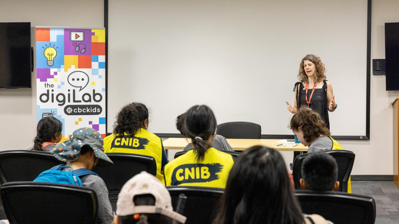 Au Maison de Radio-Canada à Toronto, un membre du personnel de la  Radio-Canada fait une présentation à un groupe de participants au Camp Connect d’INCA. À côté du présentateur se trouve une bannière sur laquelle est inscrit « The digiLab – CBC Kids ».
