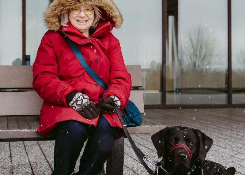 Cheri, vêtue d'une parka rouge à capuche, est assise sur un banc en bois, accompagnée de Sassy, son chien-guide labrador noir d'INCA, par une journée enneigée.