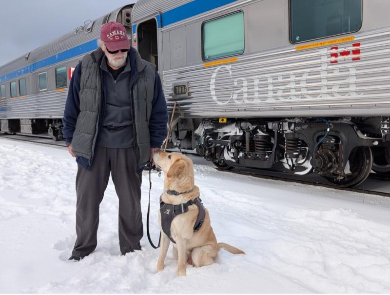 Dan se tient dans la neige aux côtés de son chien-guide, Sparky, un labrador blond, qui est assis, son harnais aux épaules, et lève les yeux vers lui. Un train de voyageurs VIA Rail est à l’arrêt sur les voies derrière eux.