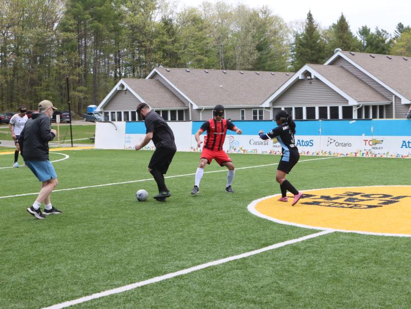 Des joueurs de soccer aveugles, aux teints variés, vêtus de tenues de sport et portant un masque sur les yeux, courent après un ballon sur le terrain de soccer accessible au camp Lake Joe d’INCA.