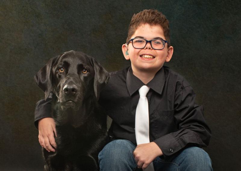 Un enfant portant des lunettes et un t-shirt noir avec une cravate blanche est assis à côté d'un chien noir et sourit.
