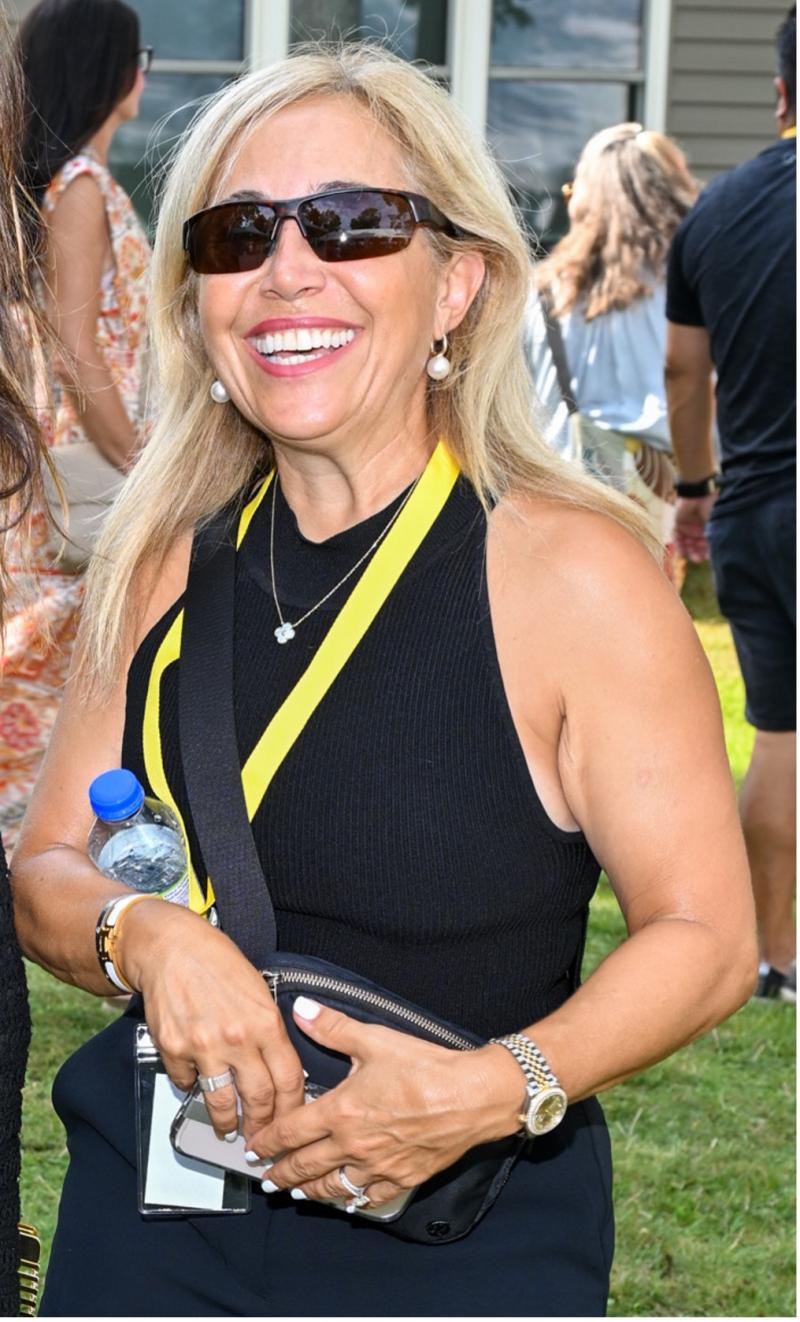 Frances est en plein air, avec un groupe de personnes qui s’affairent derrière elle. Elle porte des lunettes de soleil et sourit à la caméra.