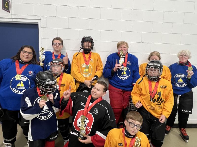 Un groupe d'enfants et d'adolescents, tous à la peau claire, pose dans une patinoire, vêtus d'équipements de hockey, notamment des maillots du camp Lake Joe d'INCA.