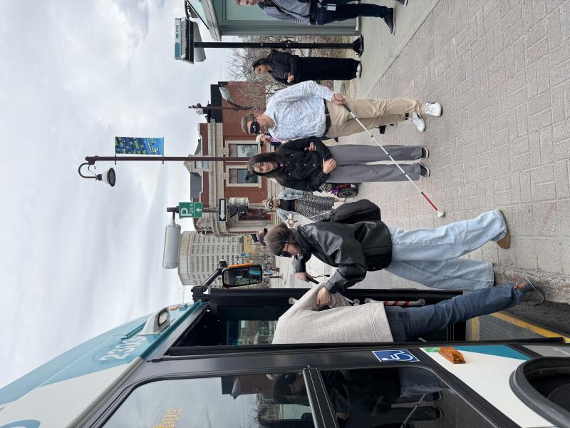Des personnes montent dans un bus urbain à un arrêt situé sur le trottoir, tandis que d'autres, qui ont les yeux bandés et utilisent des cannes blanches dans le cadre d'une simulation, attendent à proximité.